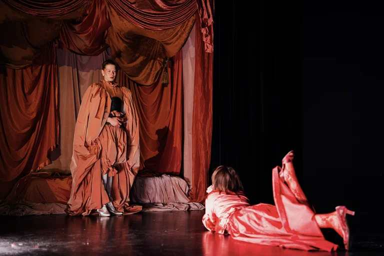 Zwei Schauspieler in orangen Kostümen führen auf einer Bühne vor roten Vorhängen ein Theaterstück auf. Dramatische Pose.