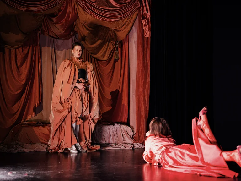 Zwei Schauspieler in orangen Kostümen führen auf einer Bühne vor roten Vorhängen ein Theaterstück auf. Dramatische Pose.