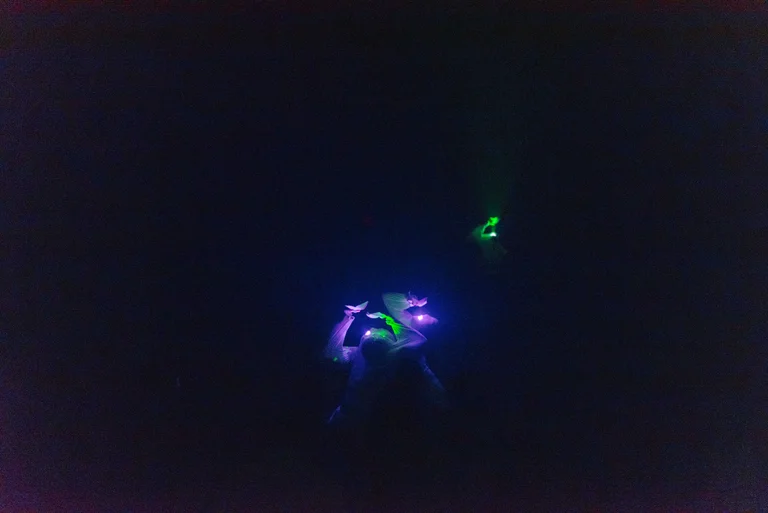 Eine Person tanzt in der Dunkelheit mit fluoreszierenden Lichtern in Blau und Grün, was eine futuristische Atmosphäre schafft.