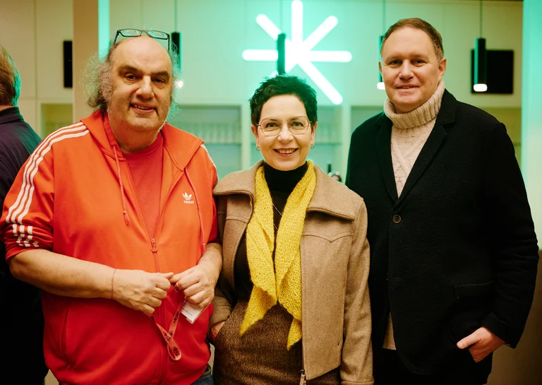 Drei Personen stehen lächelnd nebeneinander in einem beleuchteten Raum mit modernem Design. Eine Person trägt einen roten Trainingsanzug.