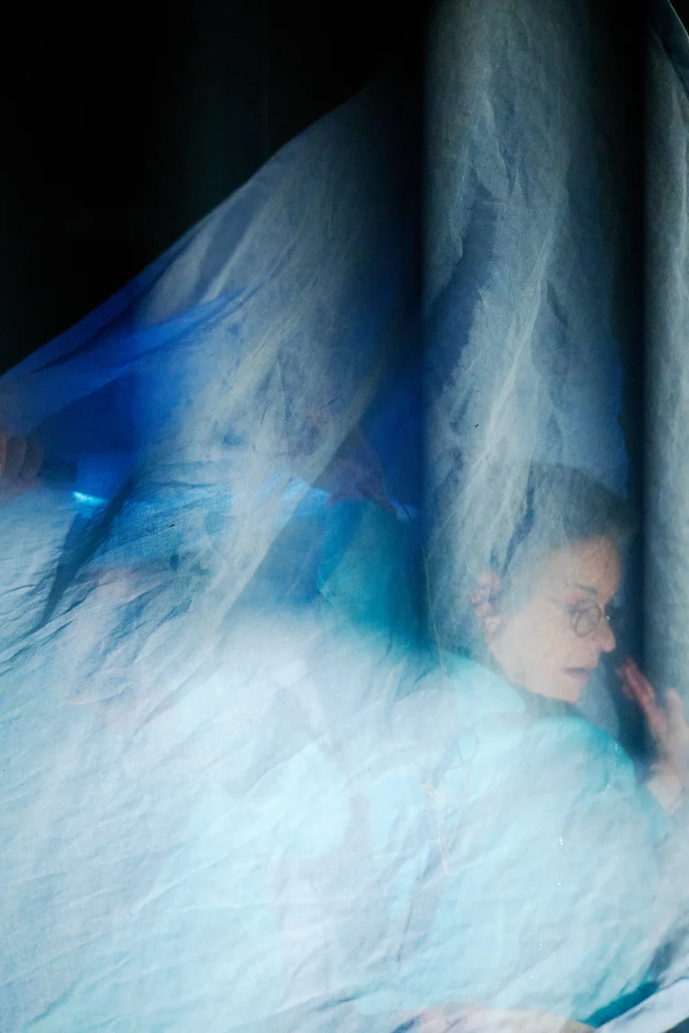 Eine verschwommene Szene mit einer Person, die hinter einem blauen, durchscheinenden Stoff ist. Die Atmosphäre wirkt traumhaft und geheimnisvoll.
