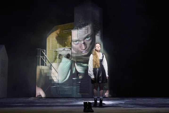 An actor stands on stage in historical costume. In the background, there's a large projected image of a pensive face.