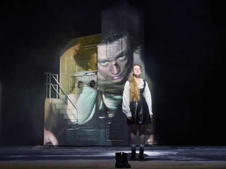 Ein Schauspieler steht auf einer Bühne in historischer Kleidung. Im Hintergrund ist ein großes projiziertes Bild mit einem nachdenklichen Gesicht.