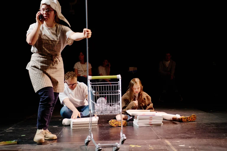 Szene aus einem Theaterstück mit einer Person im Kostüm, mit Einkaufswagen und zwei Personen, die auf dem Boden arbeiten.
