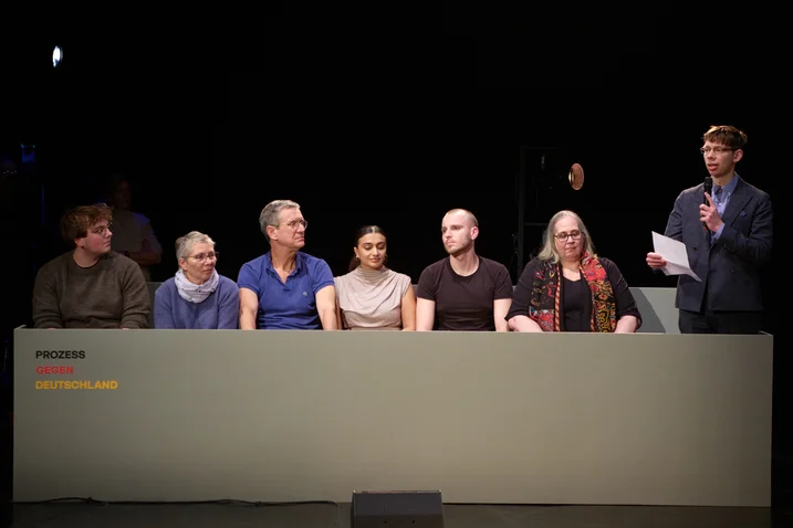 Eine Gruppe von sechs Personen sitzt an einem Tisch auf einer Bühne. Eine Person steht und liest von einem Blatt Papier.