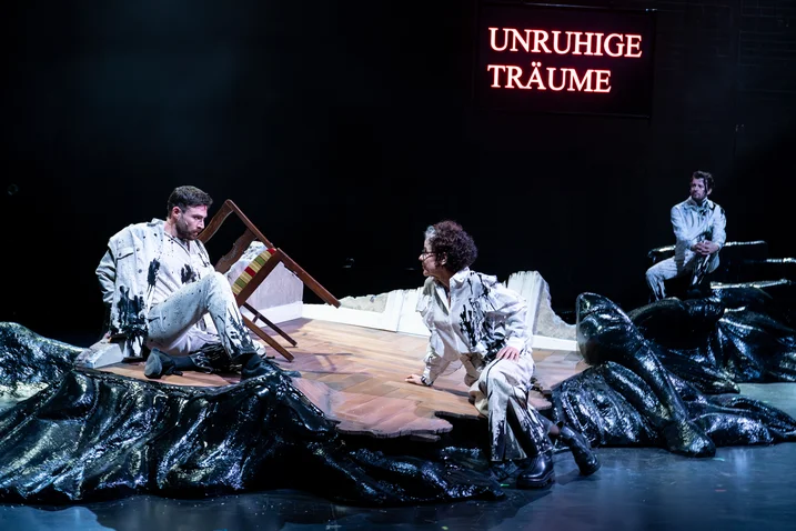 Theatre scene with three people in white outfits on stage with a black backdrop. The text "UNRUHIGE TRÄUME" glows red.