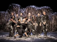 A group of people in black costumes stands in front of a dark, rugged backdrop. They pose in theatrical positions.