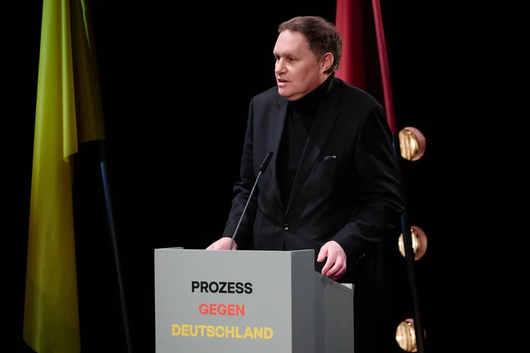 Eine Person steht an einem Rednerpult mit der Aufschrift "Prozess gegen Deutschland", im Hintergrund sind Flaggen zu sehen.