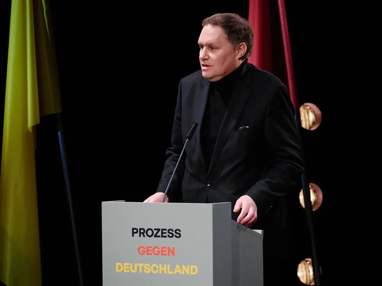 Eine Person steht an einem Rednerpult mit der Aufschrift "Prozess gegen Deutschland", im Hintergrund sind Flaggen zu sehen.