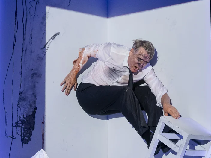 Ein Darsteller in einem weißen Hemd und schwarzer Hose klettert in einer künstlerischen Pose über einen weißen Stuhl in einer Ecke.