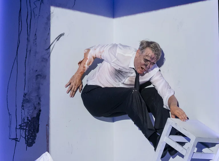 Ein Darsteller in einem weißen Hemd und schwarzer Hose klettert in einer künstlerischen Pose über einen weißen Stuhl in einer Ecke.