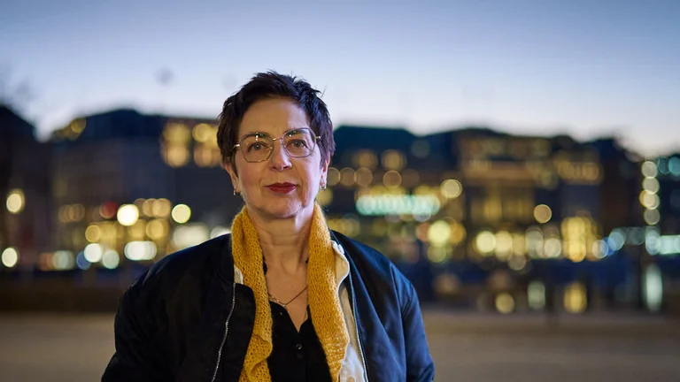 Eine Person steht in einer städtischen Umgebung bei Abenddämmerung. Im Hintergrund sind verschwommene Lichter zu sehen.