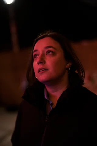 Eine Frau steht im Dunkeln, das Gesicht sanft von rotem Licht beleuchtet. Sie schaut nachdenklich in die Ferne.