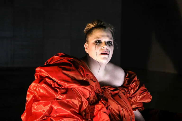 Eine Frau in einem dramatisch beleuchteten Raum trägt ein auffälliges rotes Kleid. Ihr Ausdruck wirkt intensiv und emotional.