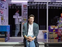 A man sits thoughtfully in front of a brightly lit kiosk with stocked shelves. Another person is visible in the background.