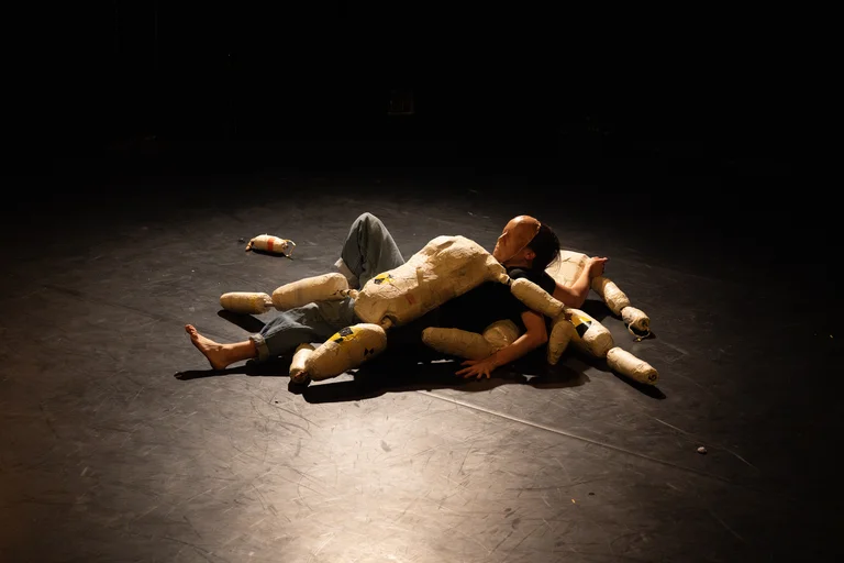 Ein Performer liegt auf einer dunklen Bühne, umgeben von Stoffpuppen. Die Beleuchtung ist dramatisch und konzentriert sich auf die Szene.