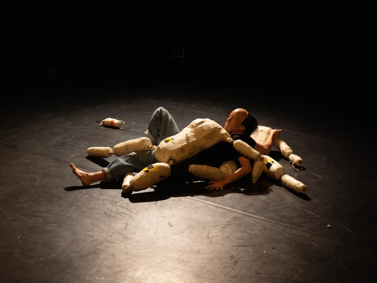 Ein Performer liegt auf einer dunklen Bühne, umgeben von Stoffpuppen. Die Beleuchtung ist dramatisch und konzentriert sich auf die Szene.