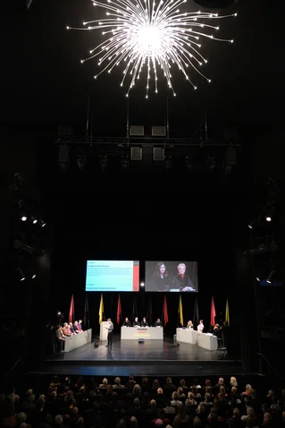 Menschen auf einer Bühne diskutieren, während ein großes Publikum zuschaut. Über ihnen leuchtet ein imposanter Kronleuchter.