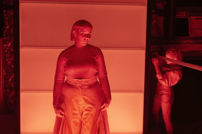 A person stands bathed in red light in front of a glowing wall. Another figure in the background holds an object.