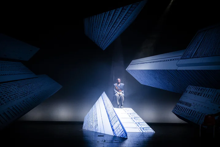 A performer stands on an illuminated stairway in an artful, abstract stage setting with large, blue geometric shapes.
