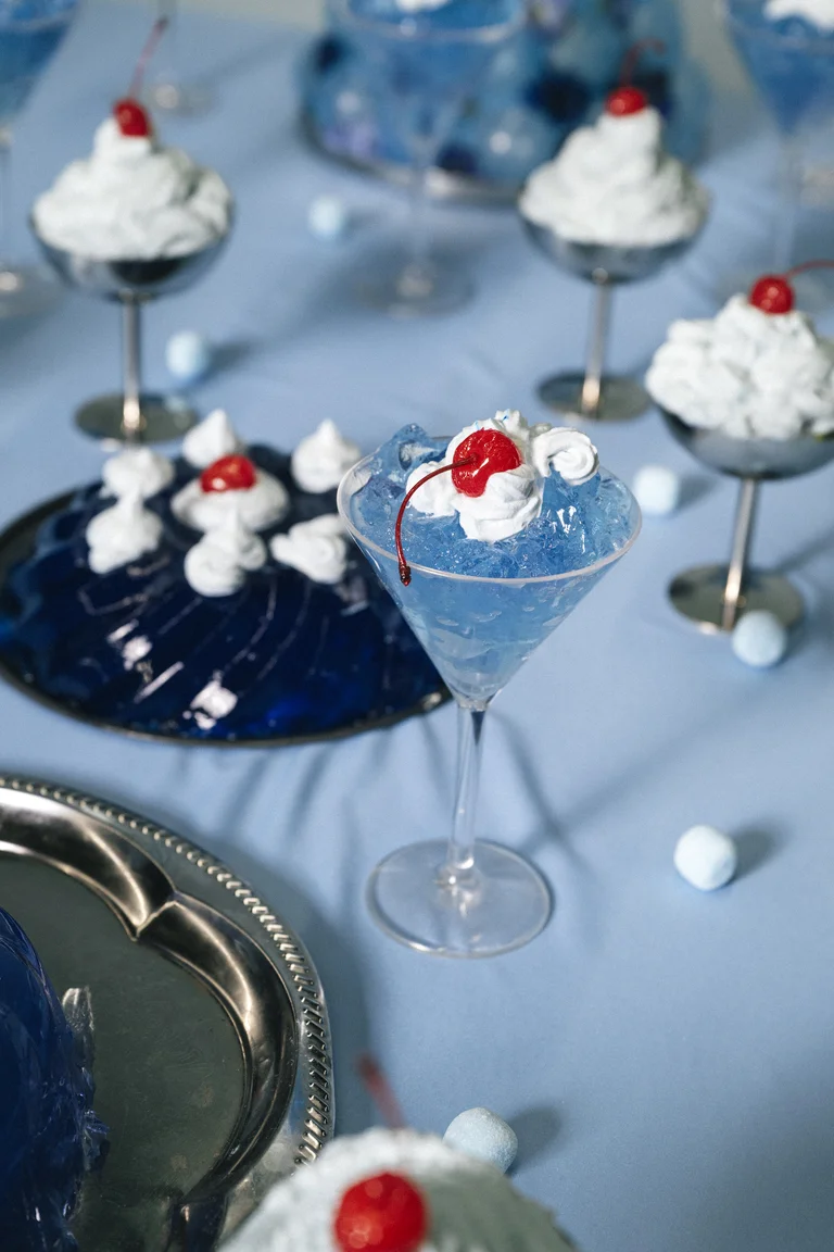 Desserts in elegant glasses on a blue background. Blue jelly with whipped cream and cherries on top, served on a tray.