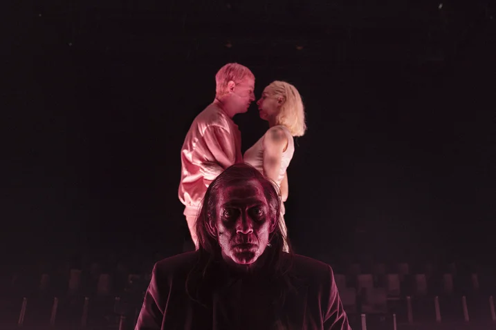 Three people on stage; two stand closely together in the background, while a third stands in front, under dramatic lighting.