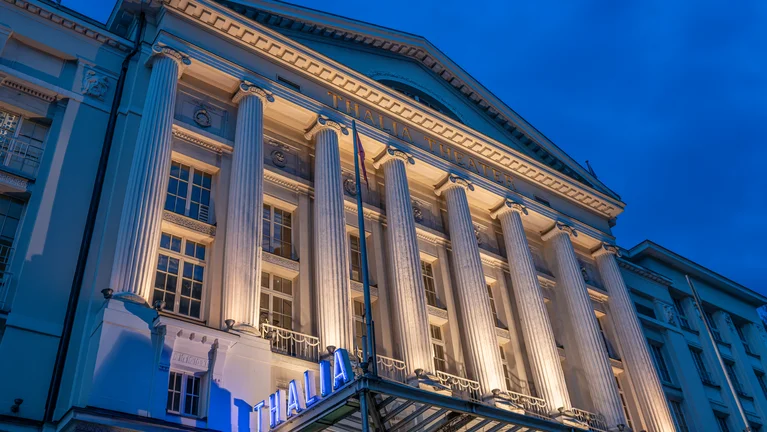 A large theater building with neoclassical columns and blue lighting, majestic in the evening.