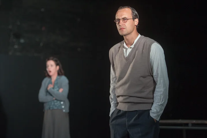 A man and a woman stand on a stage. The man wears a sweater, while the woman has her arms crossed.