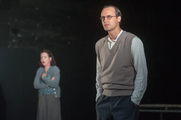 Ein Mann und eine Frau stehen auf einer Bühne. Der Mann trägt einen Pullover und die Frau hat die Arme verschränkt.