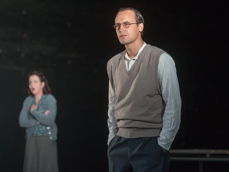 Ein Mann und eine Frau stehen auf einer Bühne. Der Mann trägt einen Pullover und die Frau hat die Arme verschränkt.