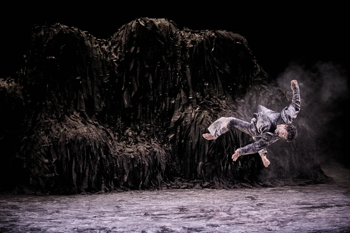 A dancer in a gray costume gracefully leaps mid-air against a dark, mysterious backdrop. His movement is acrobatic and dramatic.