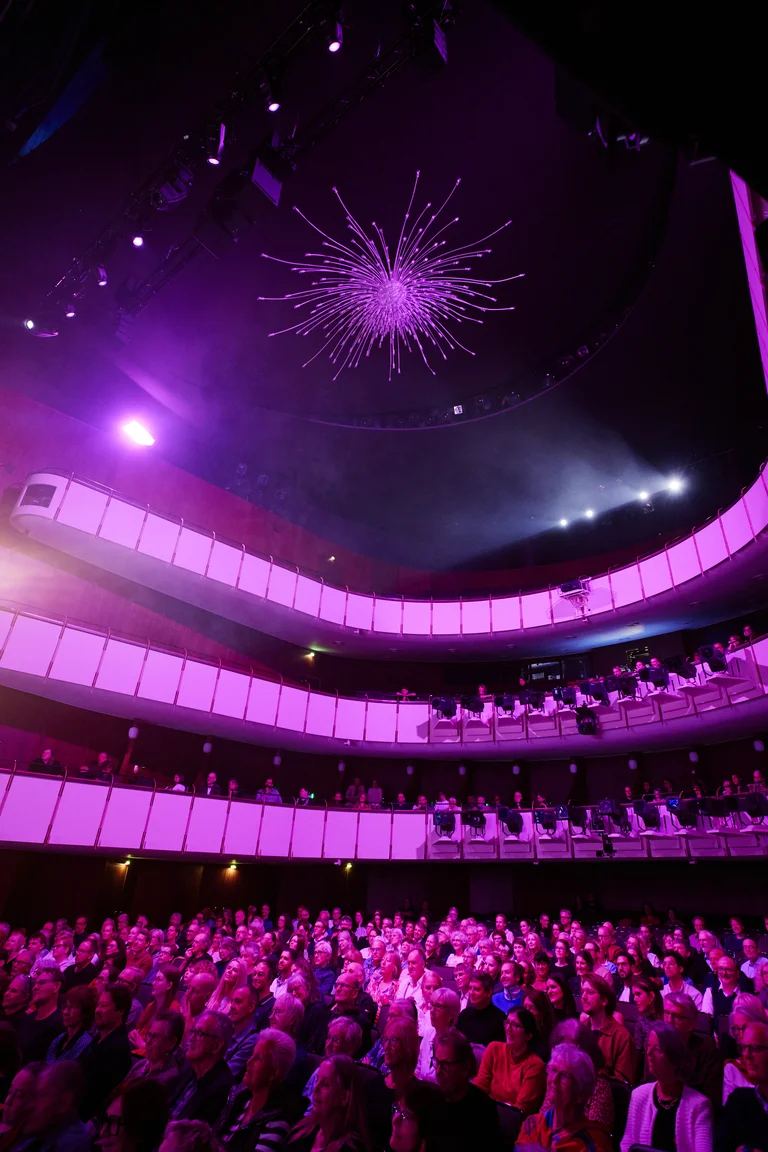 Ein mehrstöckiges Theater mit Zuschauern auf allen Ebenen, beleuchtet von violettem Licht. Eine sternförmige Dekoration hängt von der Decke.