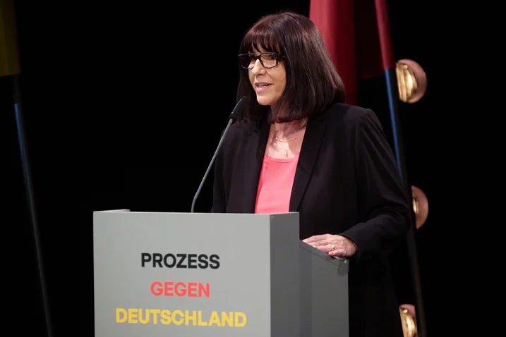 Eine Frau spricht auf einer Bühne an einem Podium mit der Aufschrift "Prozess gegen Deutschland". Hintergrund mit Flaggen.