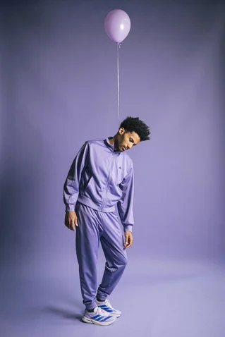 A man in a purple tracksuit stands in a studio. A purple balloon floats above his head.