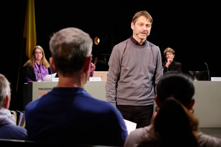 Ein Mann steht vor einem Publikum, während einige Personen an einem Tisch im Hintergrund sitzen. Die Atmosphäre ist konzentriert.