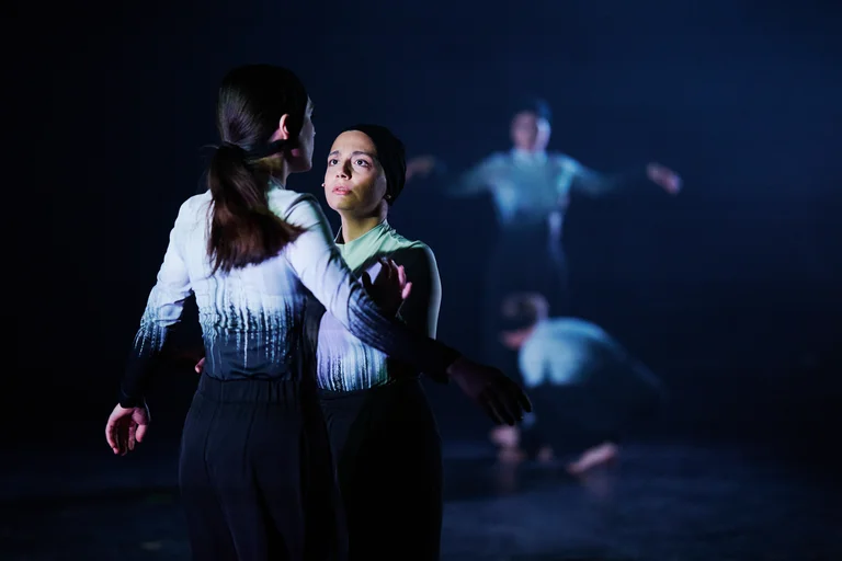 Zwei Tänzerinnen in Schwarz-Weiß-Outfits auf dunkler Bühne, im Hintergrund weitere Tänzer. Intensives, ausdrucksstarkes Modern Dance-Performance.
