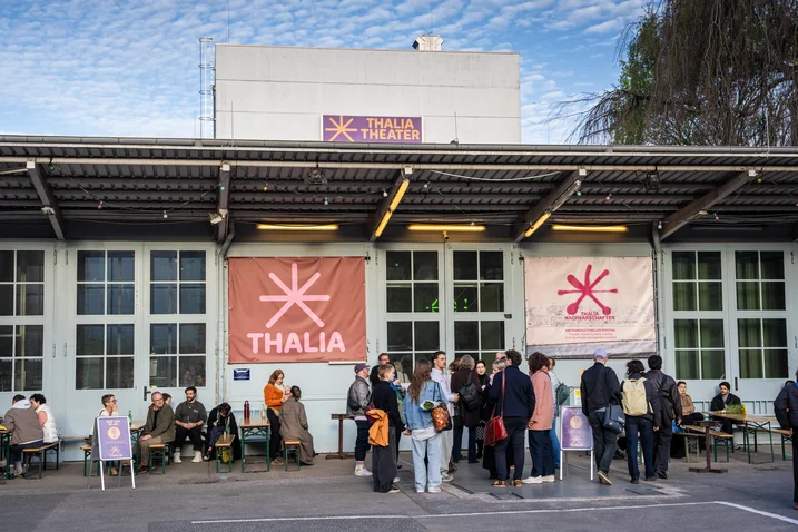 Eine Menschengruppe steht vor dem Thalia Theater mit Bannern. Der Himmel ist bewölkt und der Tag scheint kühl zu sein.