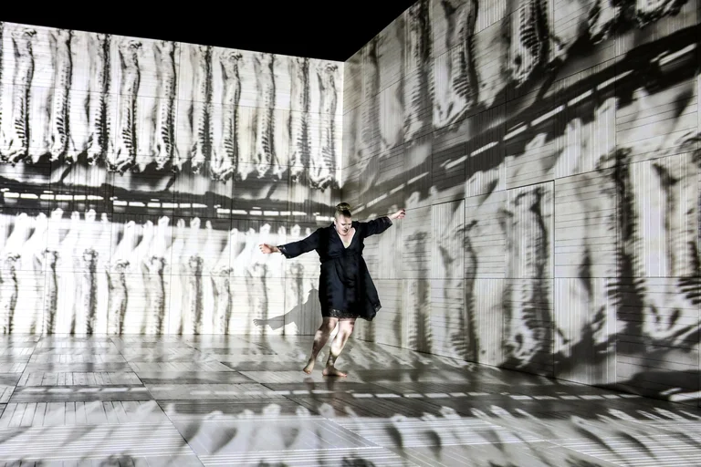 Eine Person tanzt in einem Raum mit gestreiften und texturierten Wänden, die Schatten und Lichtspiele erzeugen. Kunstvolle Inszenierung.