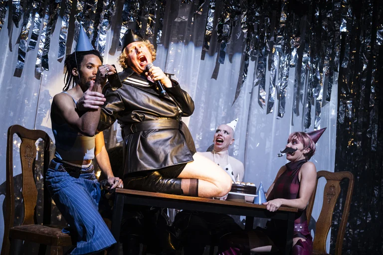 Menschen bei einer feierlichen Darbietung, ein Mann steht auf einem Tisch und singt ins Mikrofon, umgeben von Partygästen.