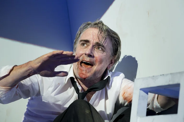 Ein Schauspieler in einem intensiven Moment einer Theateraufführung. Er trägt Hemd und Krawatte und scheint emotional erregt zu sein.