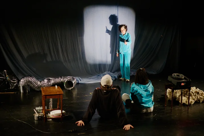 Eine Person in blauem Outfit führt auf einer Bühne eine Darbietung vor, während zwei Zuschauer von hinten betrachtet werden.