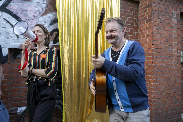 Ein Mann steht neben einer Frau mit Megafon vor einem goldenen Vorhang und einem Backstein-Gebäude. Er hält eine Gitarre.