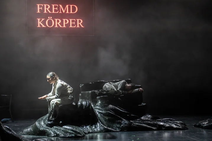 A person sits on a stage with a dark backdrop. The words "Fremd Körper" illuminate in the background.