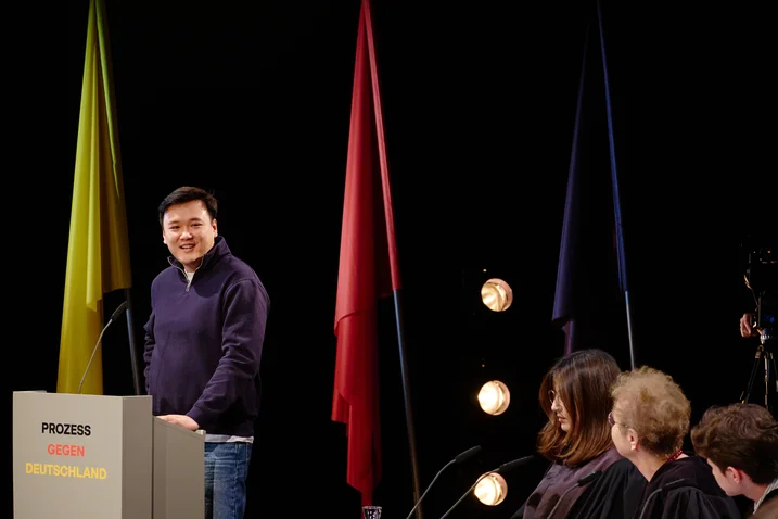 Ein Mann steht an einem Podium. Im Hintergrund sind bunte Fahnen und Scheinwerfer zu sehen.