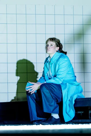 A person in a blue coat sits thoughtfully in front of a tiled wall. A shadow is visible.