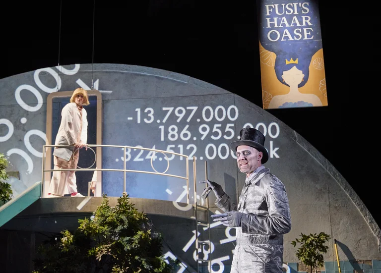 Two people on stage with projected numbers in the background. A man in a silver suit stands centrally. Banner reads "Fusi's Hair Oasis."