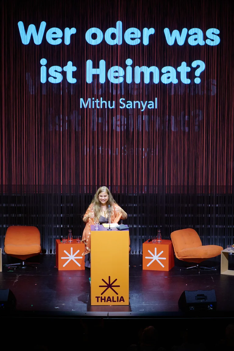 Eine Person steht an einem Rednerpult auf einer Bühne. Im Hintergrund steht "Wer oder was ist Heimat?" und "Mithu Sanyal".