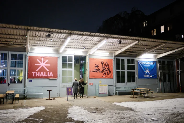 Außenansicht eines Gebäudes bei Nacht, mit beleuchteten Schildern von Thalia, einer Veranstaltung, und einem Café oder Theater.