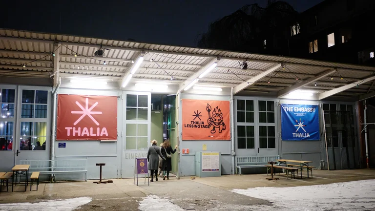 Außenansicht eines Gebäudes bei Nacht, mit beleuchteten Schildern von Thalia, einer Veranstaltung, und einem Café oder Theater.