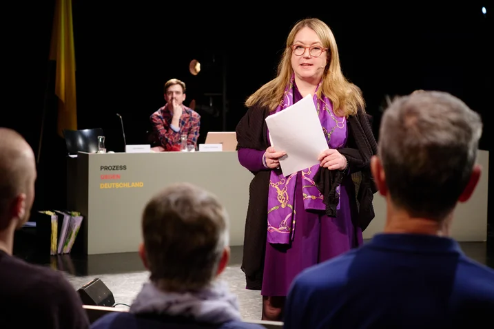 Frau hält einen Vortrag auf einer Veranstaltung, während die Zuhörer aufmerksam zuhören. Ein Tisch im Hintergrund mit einem Teilnehmer.